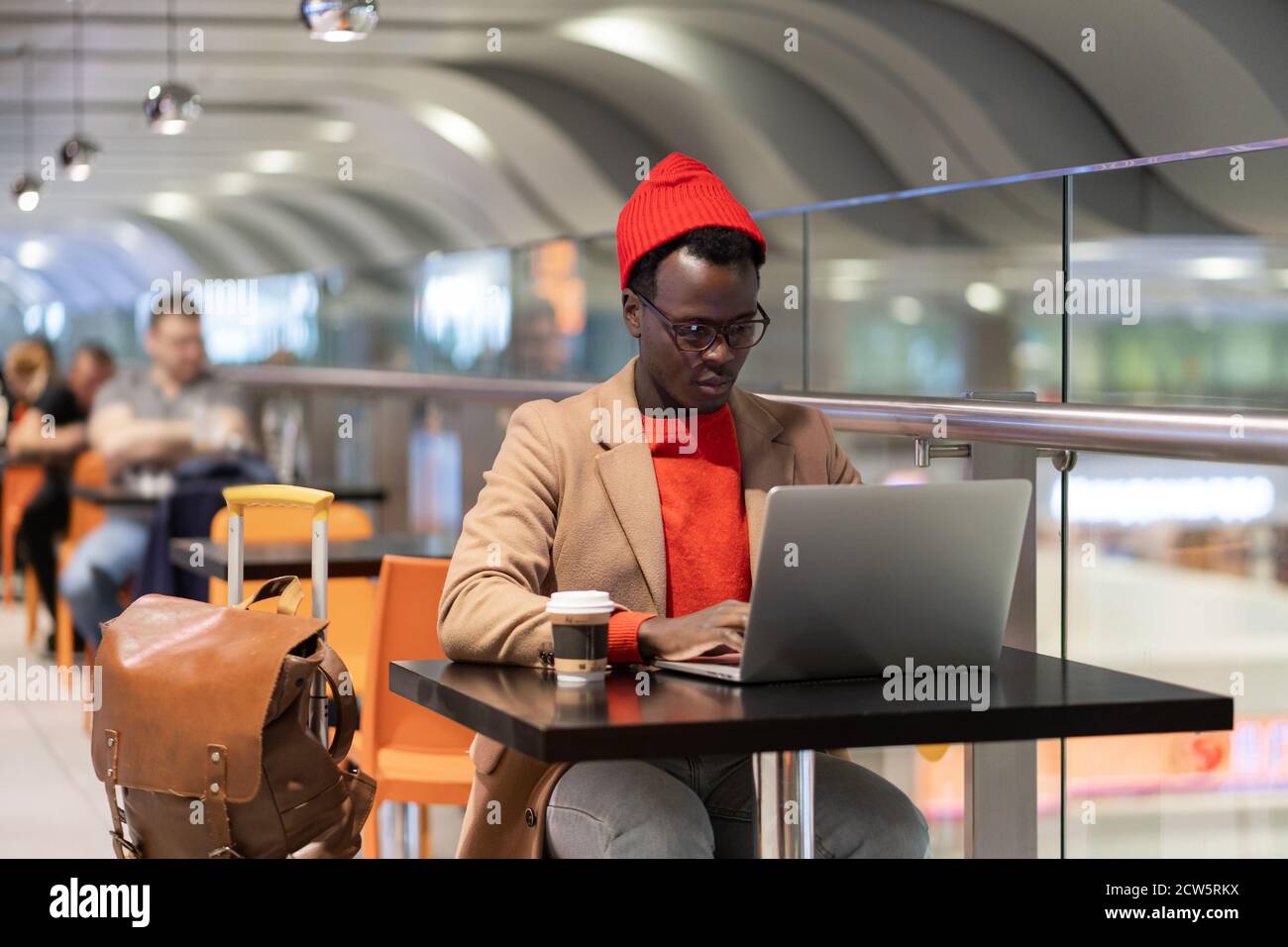 Stylish African American traveler millennial man in beige coat and red hat resting, sitting at cafe table in airport terminal, works remotely on lapto Stock Photo