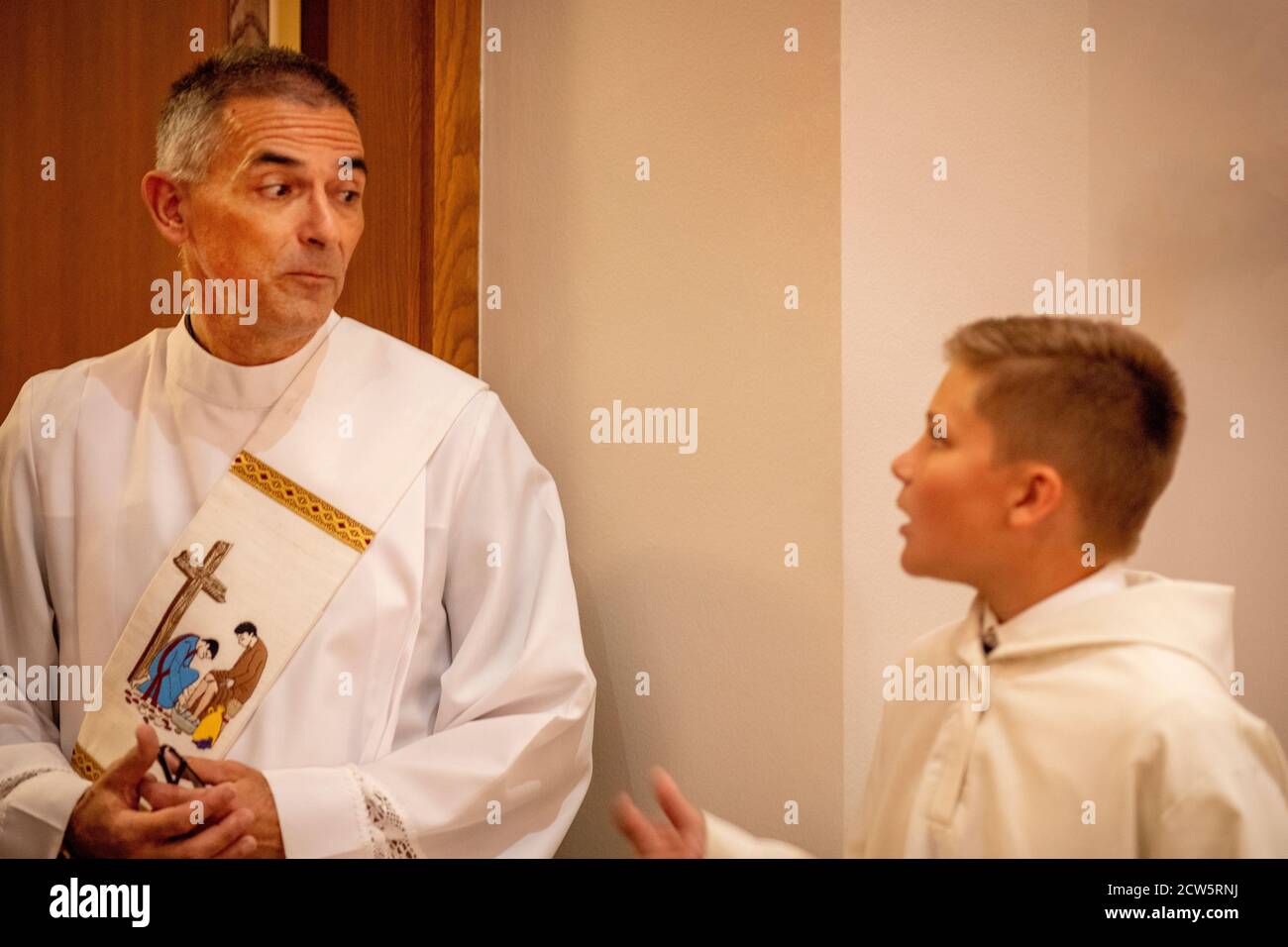 At a Southern California Catholic church, a robed deacon with a picture sash converses with a young boy altar assistant before mass. Stock Photo