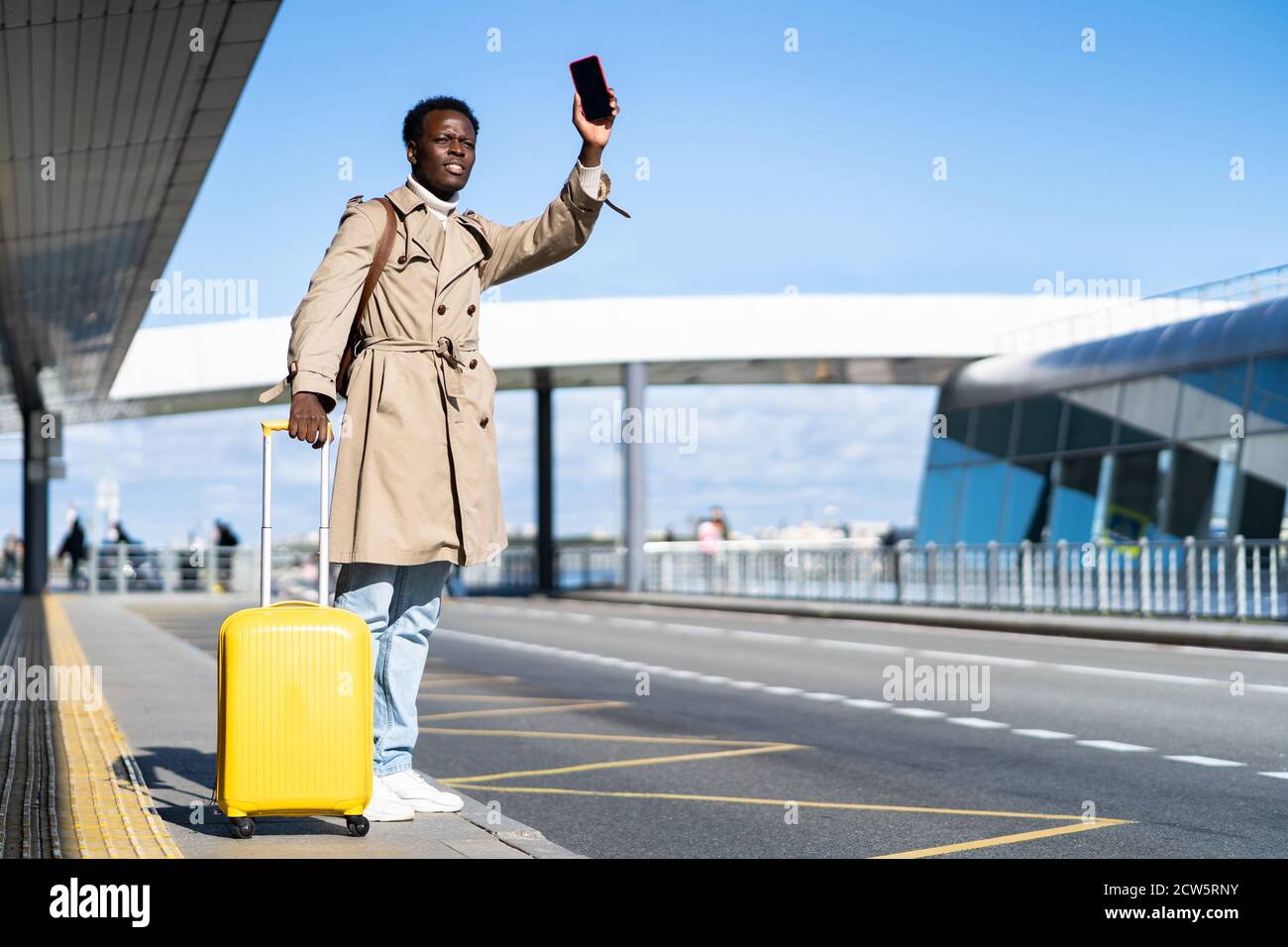 Afro-American millennial traveler man wear beige trench coat with yellow suitcase stands in airport terminal, holding phone and calling taxi, raising Stock Photo