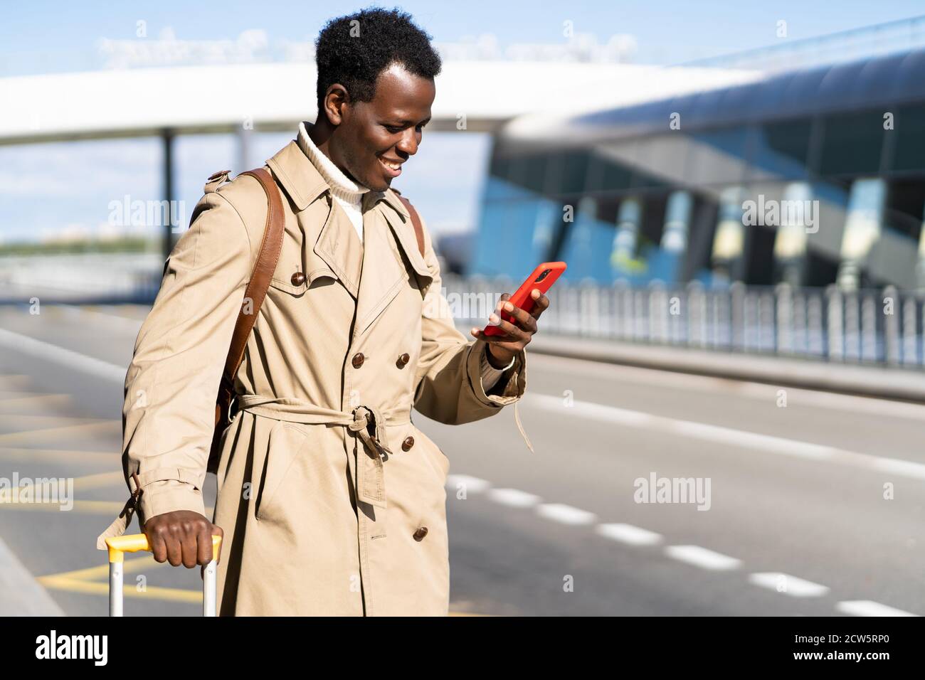 Smiling Afro-American traveler man with suitcase stands in airport terminal, using mobile application in smart phone to rent vehicle online in car sha Stock Photo