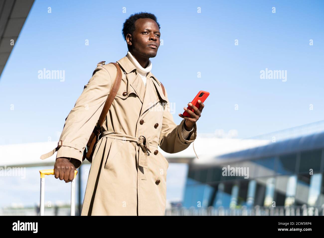 Afro-American millennial traveler man with yellow suitcase stands in airport terminal, holding phone, calling and looking for a taxi or car sharing. B Stock Photo