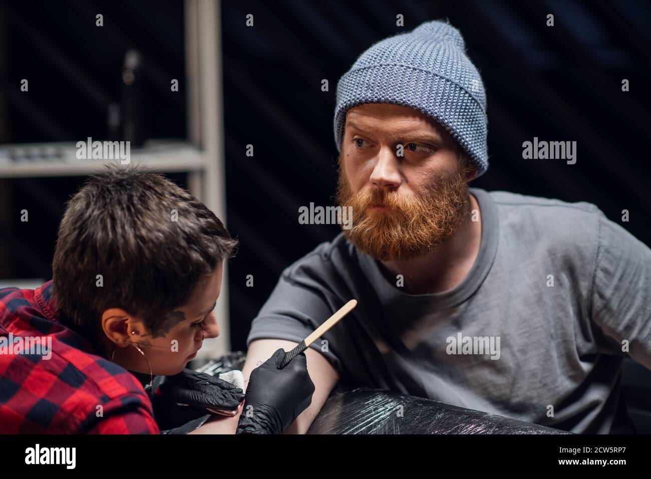 Shorthaired girl with piercings and tattoos on her face and hands the  master makes a tattoo in a handpoke way for a young man with a red beard  and a knitted hat