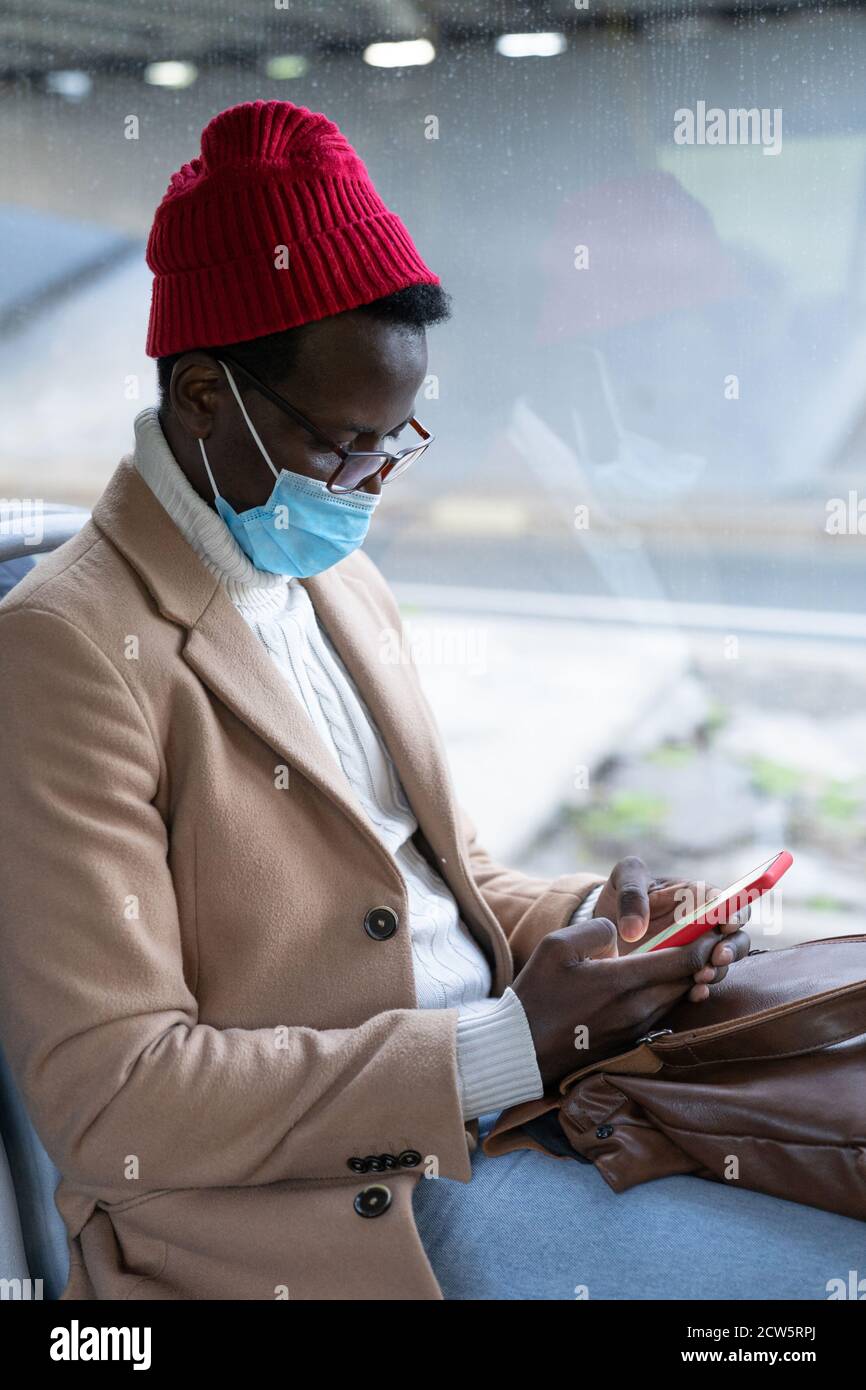 Stylish African American traveler man in beige coat, using phone, wearing mandatory face mask to protect yourself from contact with flu virus, pandemi Stock Photo