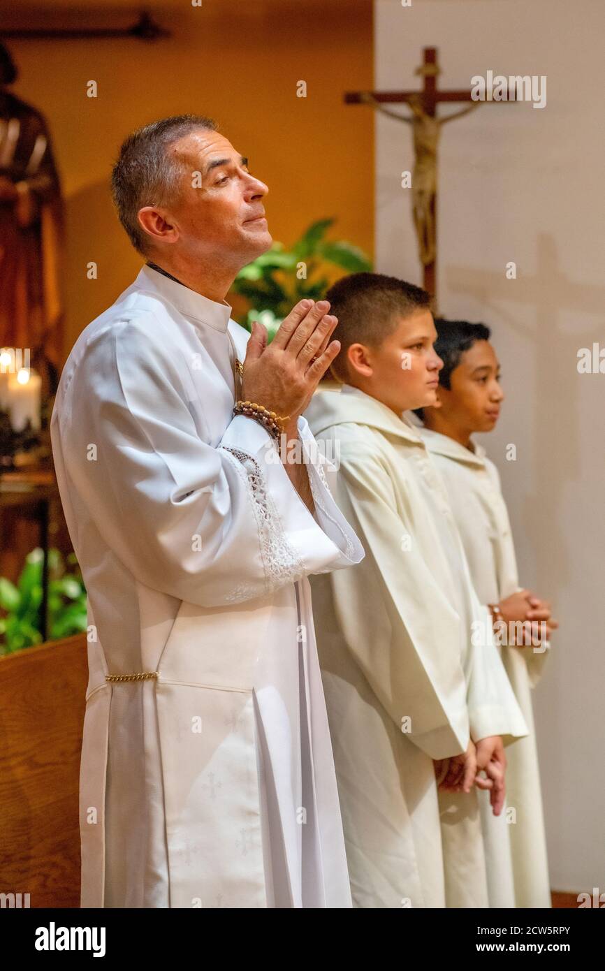 A robed deacon holds his hands in prayer along with two altar boys as they listen happily to the sermon during mass in a Southern California Catholic Stock Photo