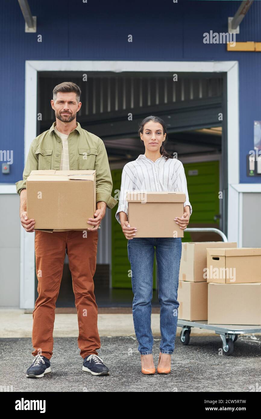 Vertical full length portrait of young couple holding boxes and looking at camera while standing by self storage facility Stock Photo