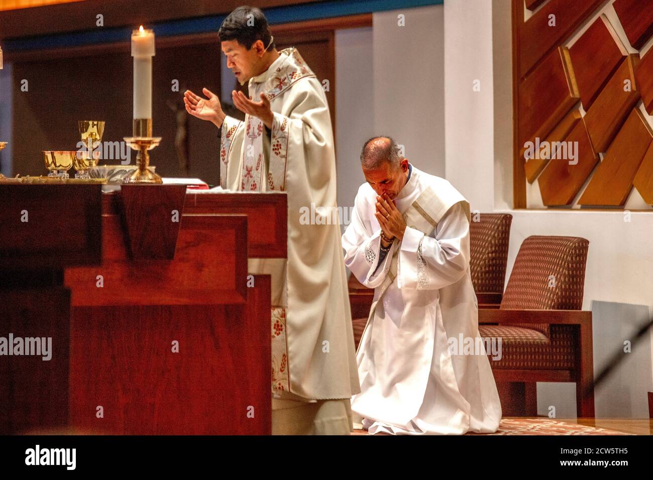 Wearing ceremonial robes, a deacon bows his head in prayer as an Asian American priest conducts mass at the altar of a Southern California Catholic ch Stock Photo