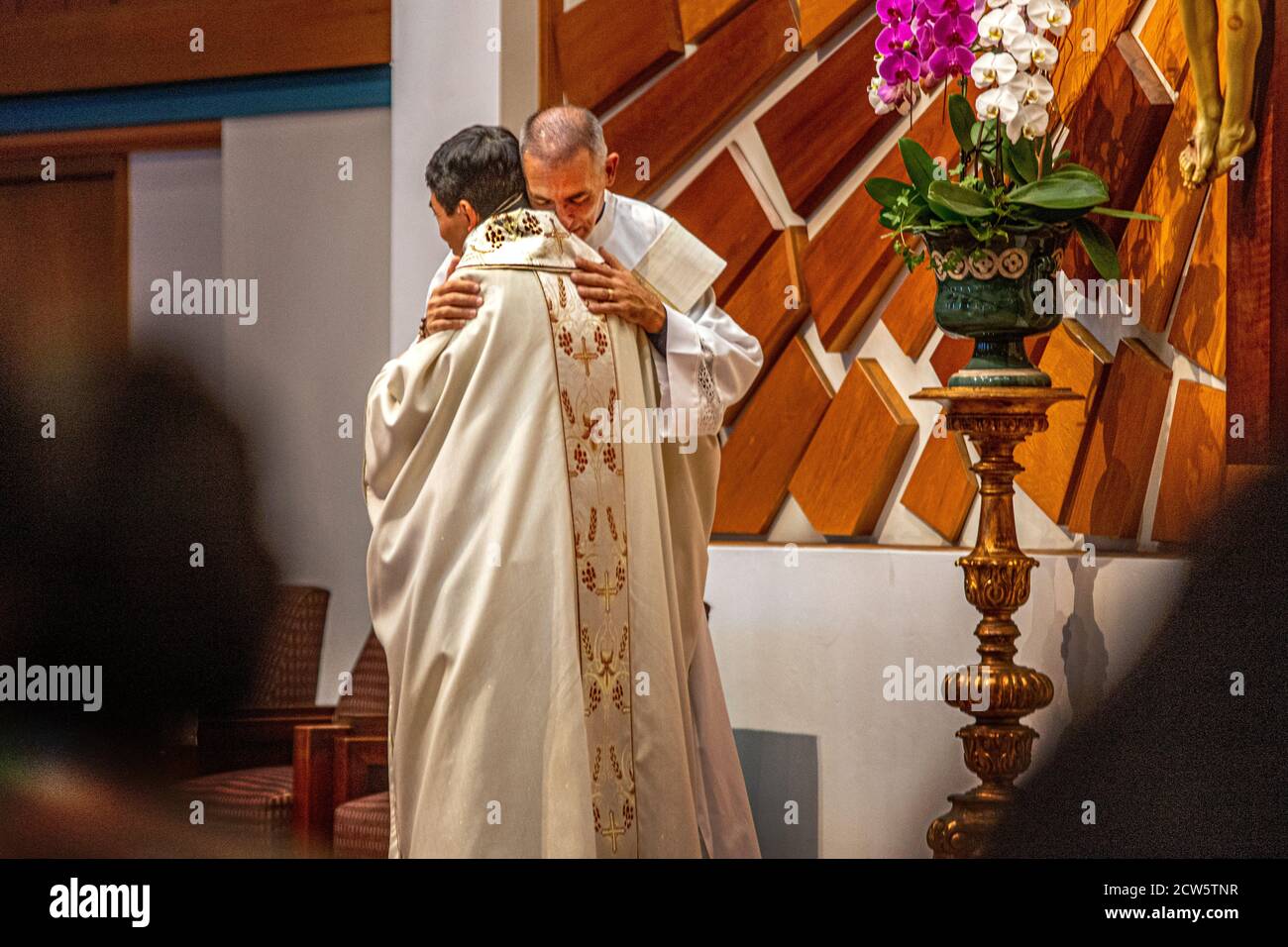 Wearing ceremonial robes, a deacon embraces an Asian American priest conducting mass at the altar of a Southern California Catholic church. Stock Photo