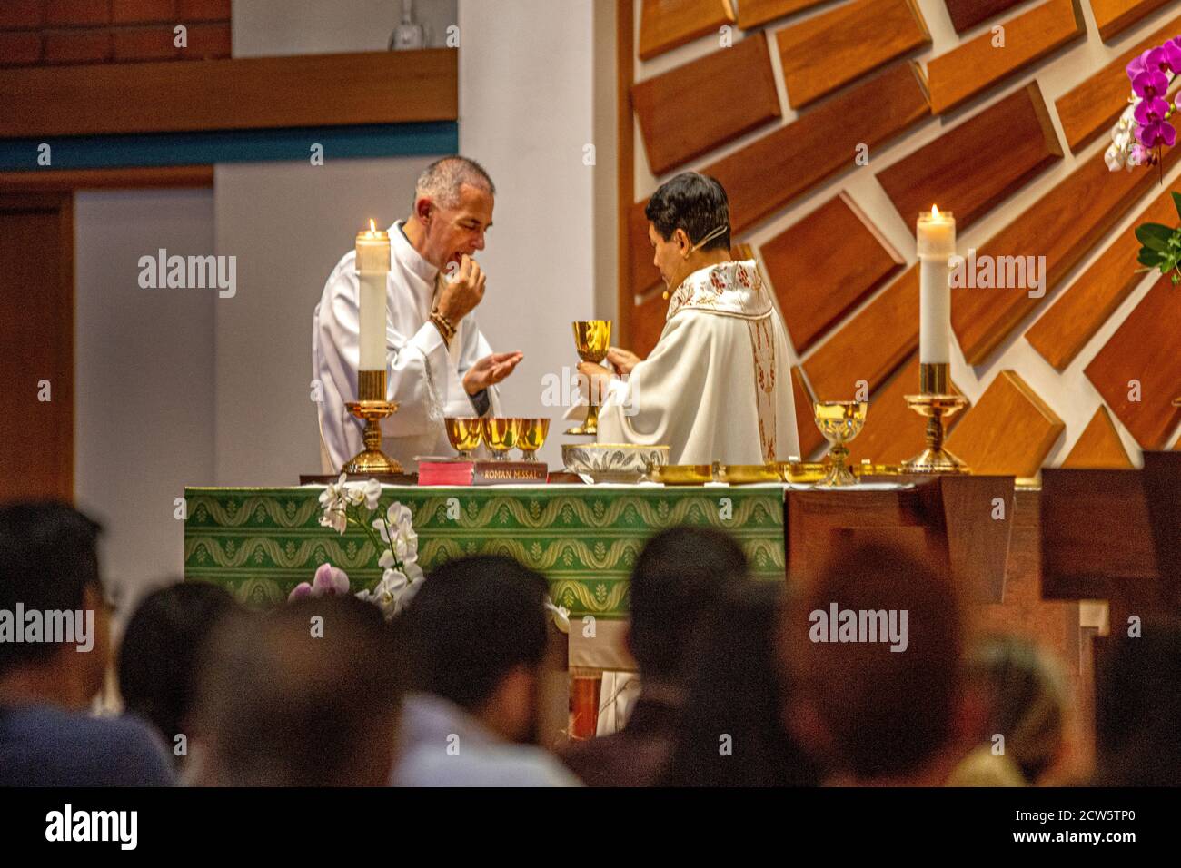 Wearing ceremonial robes, a deacon assists an Asian American priest preparing for communion during mass at the altar of a Southern California Catholic Stock Photo