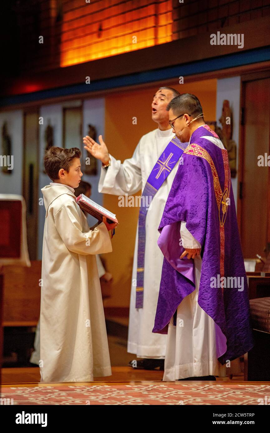 Wearing ceremonial robes, a deacon sings as an Asian American priest conducts mass at the altar of a Southern California Catholic church and an altar Stock Photo