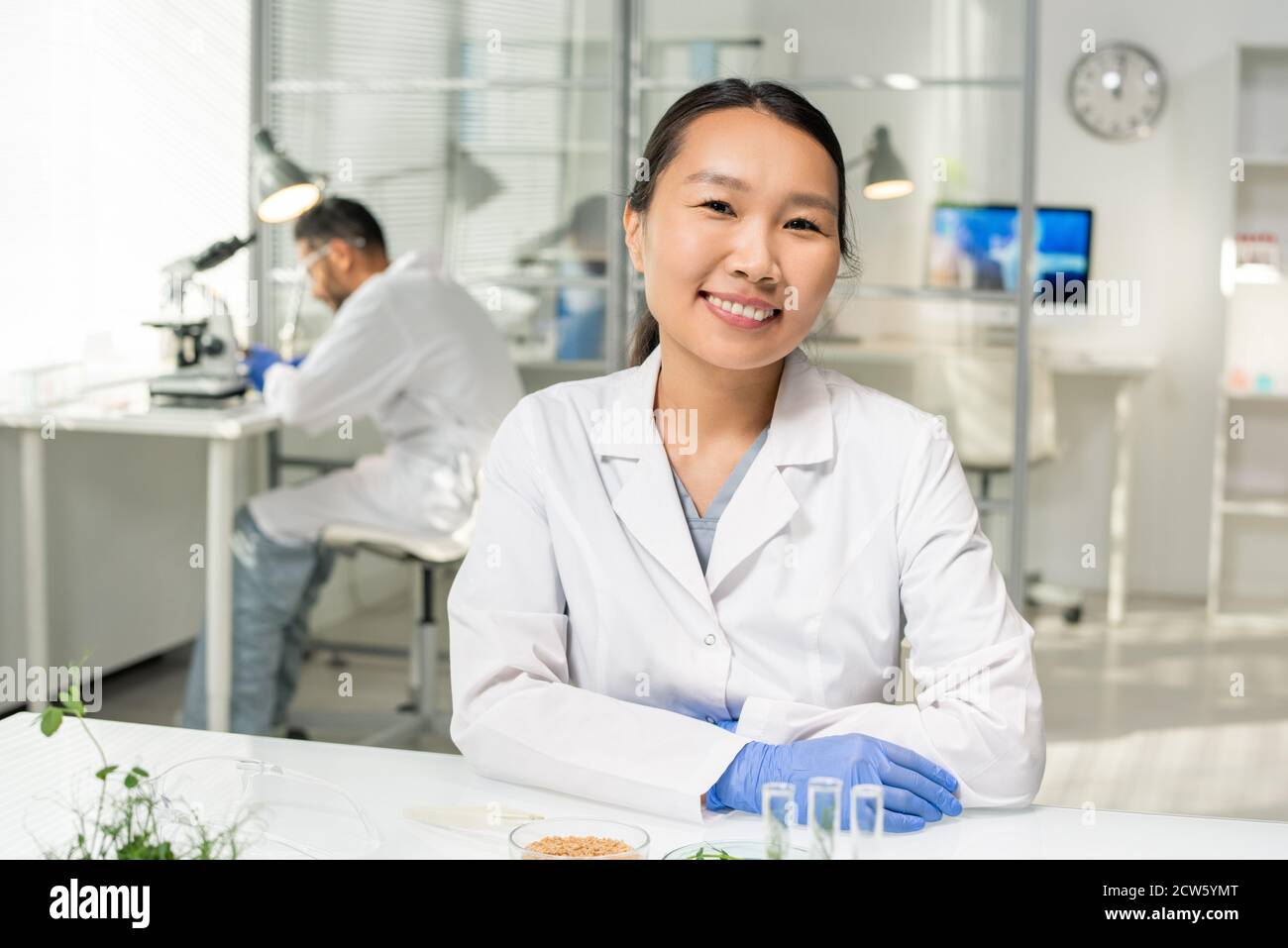 Young successful Asian female researcher in whitecoat and gloves looking at you Stock Photo