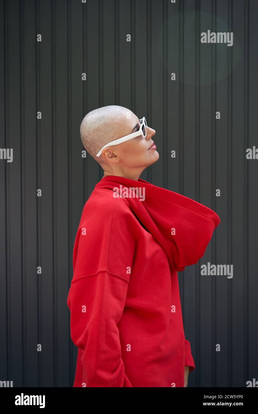Cool bald woman wearing red hoodie and sunglasses standing on background. Stock Photo