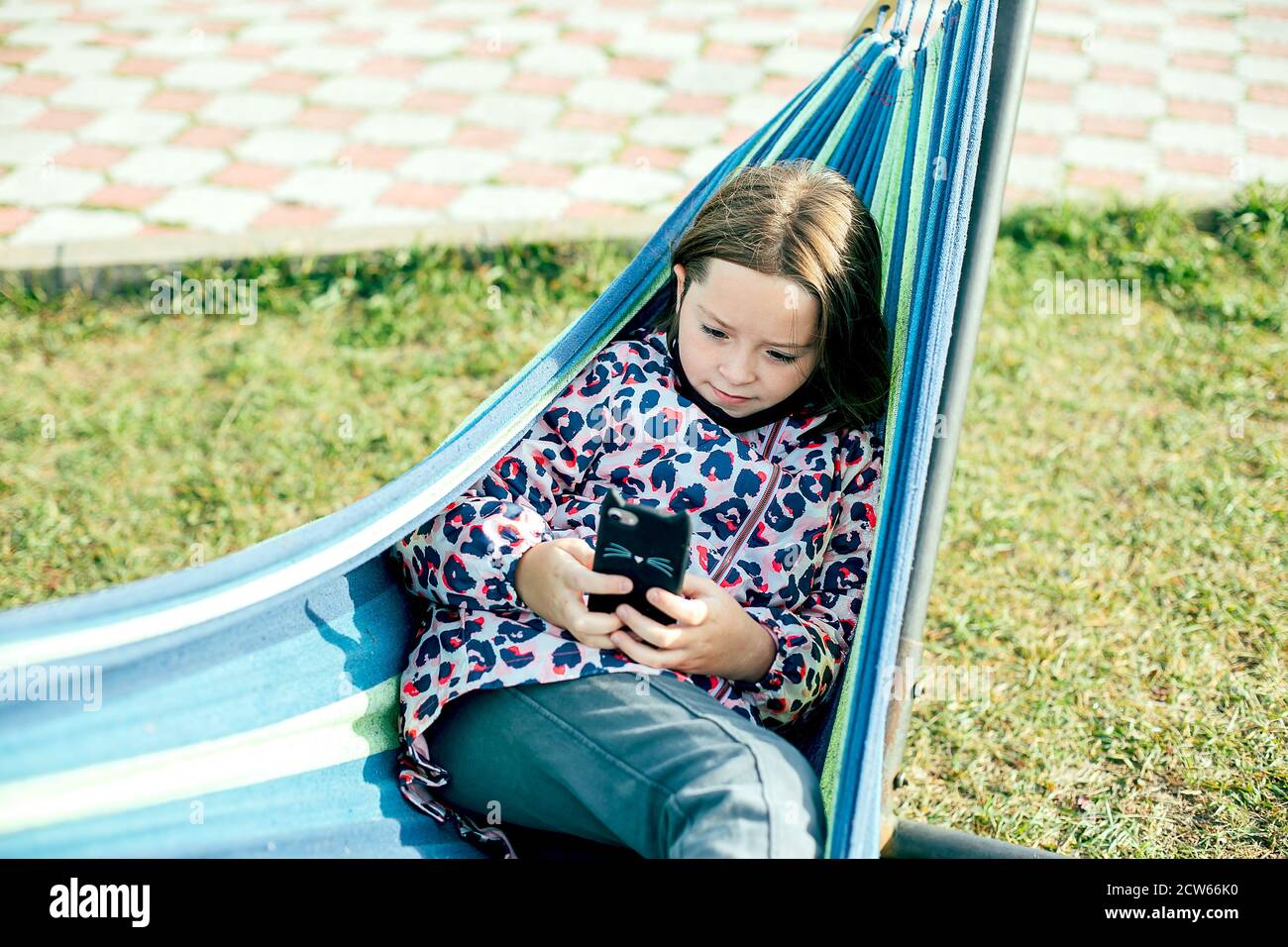 Cute caucasian girl 7-8 years old lies on hammock and using her phone on a sunny day. Stock Photo
