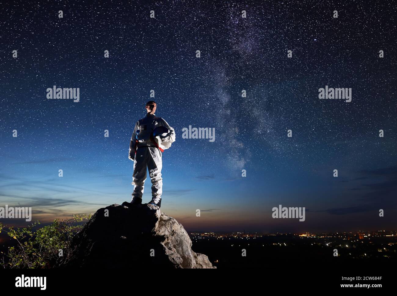 Space traveler standing on top of rocky hill and looking at beautiful sky with stars. Astronaut in white space suit holding helmet. Concept of space travel and cosmonautics. Stock Photo