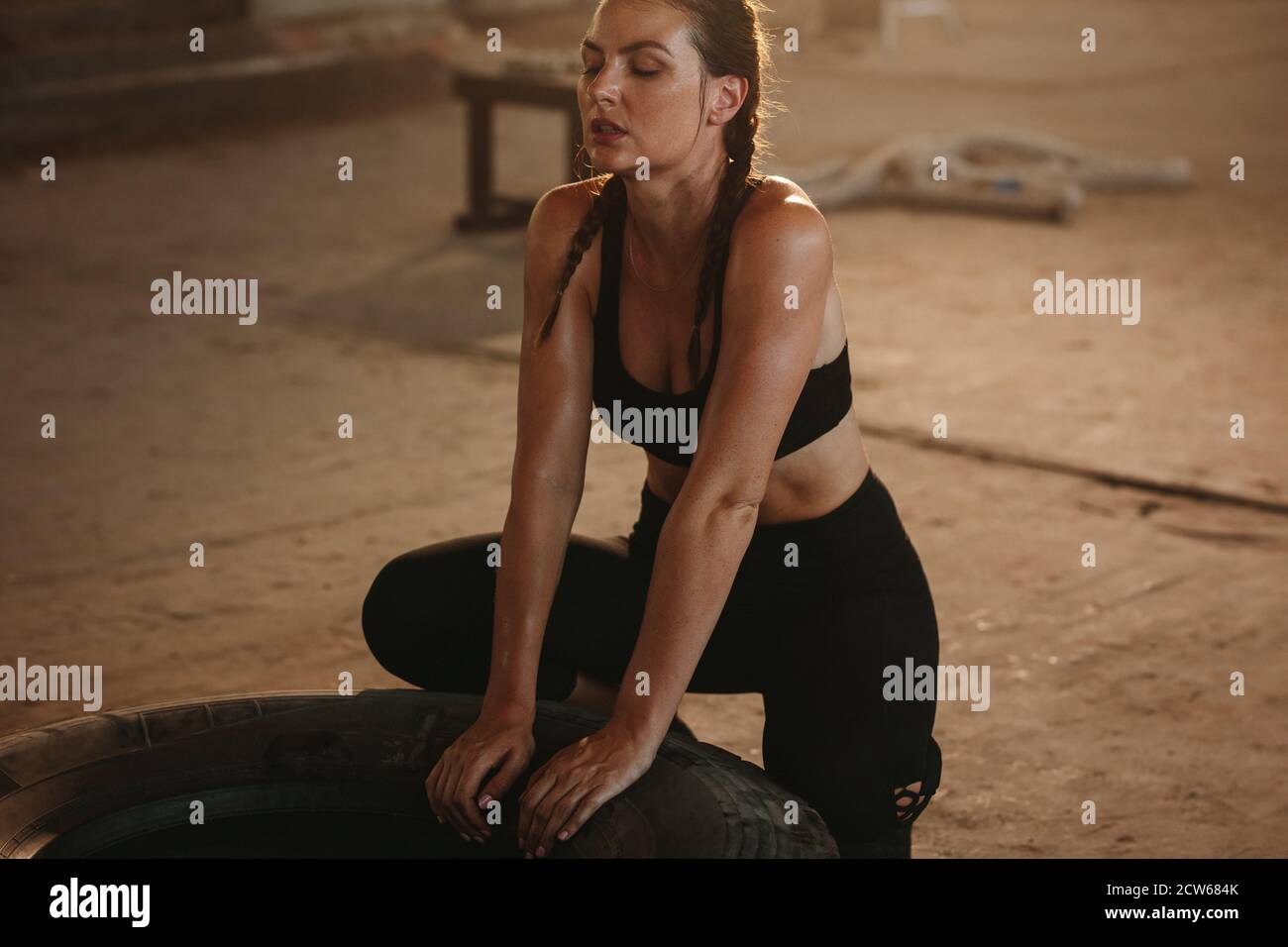 Tired and exhausted female athlete relaxing after workout. Woman taking a break after intense physical training with a tyre at cross workout space. Stock Photo
