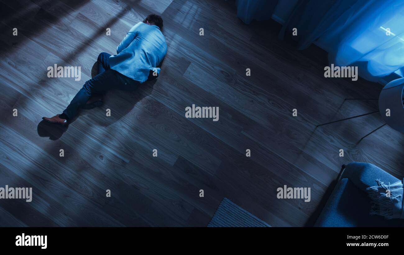 Poor Depressed Drunk Young Man is Sleaping on the Floor Near Sofa in an Apartment with Wooden Flooring. Dramatic Top View Camera Shot. Stock Photo