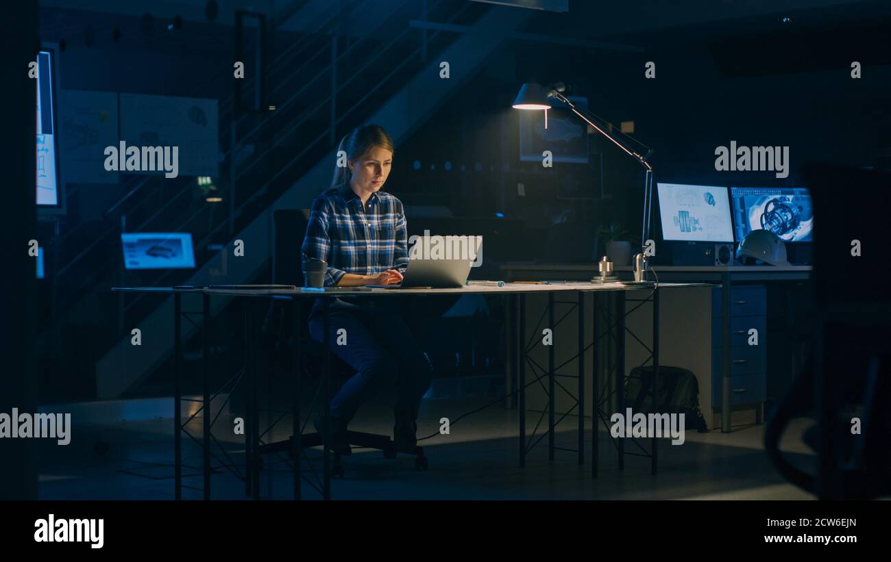 Beautiful Female Engineer Sitting at Her Desk Works on a Laptop Computer. Blueprints Lying on a Table. In the Dark Industrial Design Engineering Stock Photo