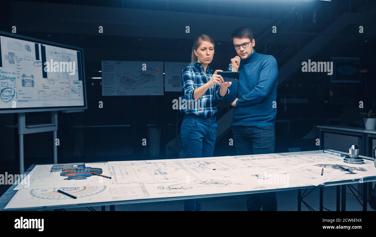 Two Young Software Development Engineers Talk and Use Smartphone with Augmented Reality for Reading and Interacting with Car and Engine Concept Stock Photo