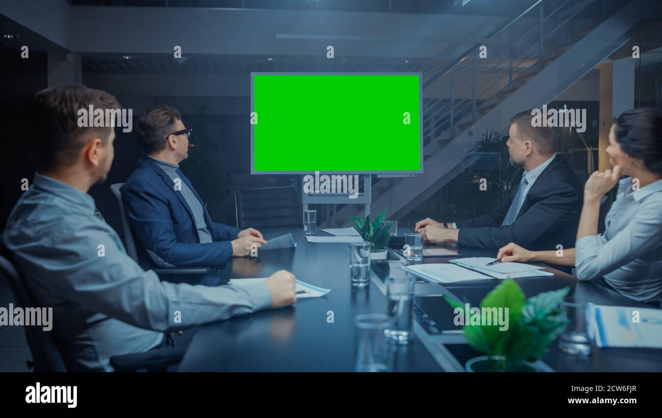 Late at Night In Corporate Meeting Room: Board of Directors, Executives and Businesspeople Sitting at Negotiations Table, Talking and Using Green Mock Stock Photo