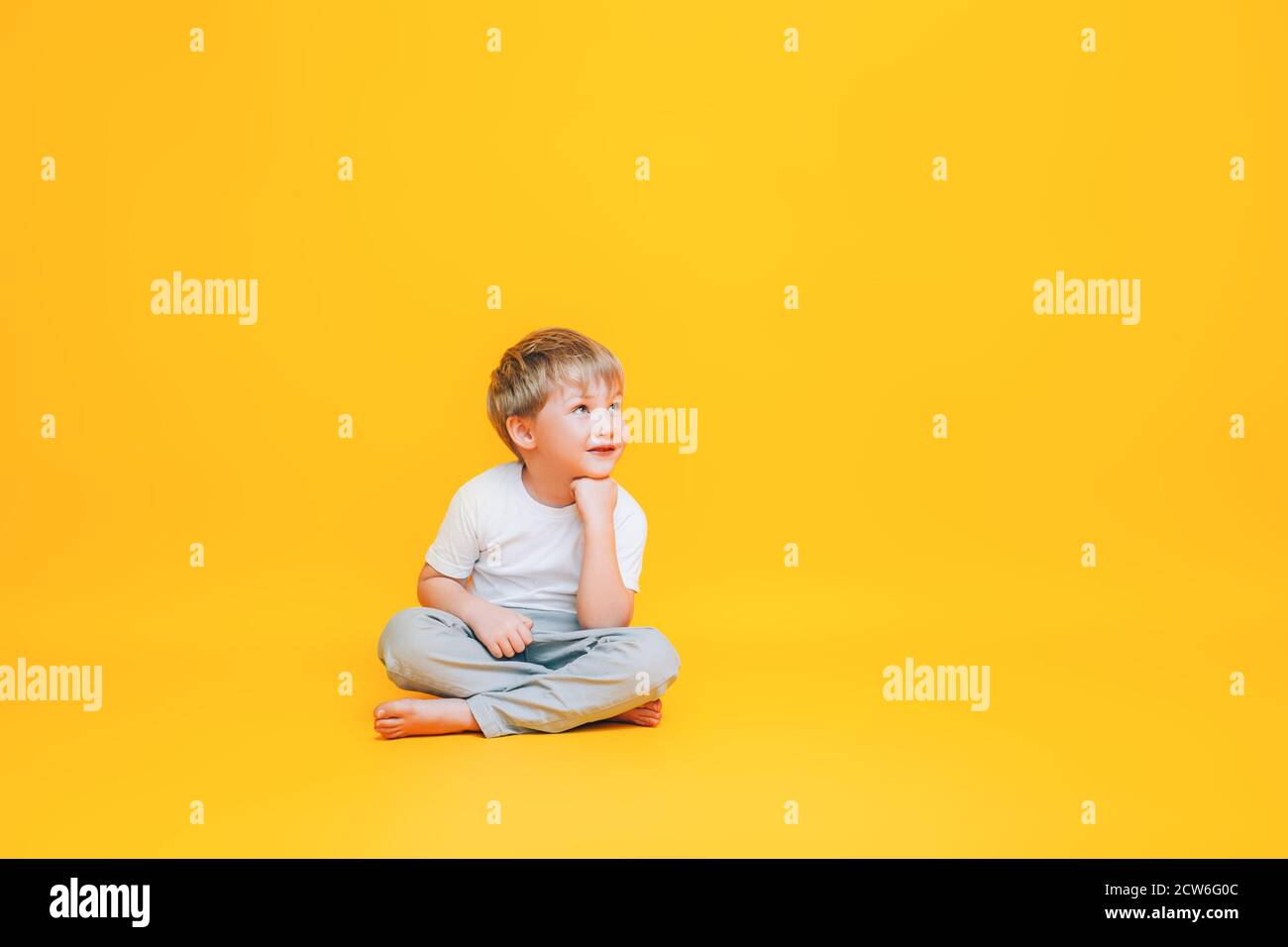 Pensive little boy in white t-shirt sitting on yellow background Stock Photo
