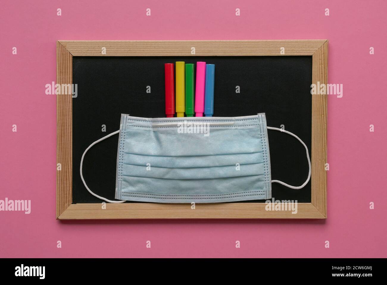 Face mask on top of colored pens and blackboard. Pink background. Stock Photo