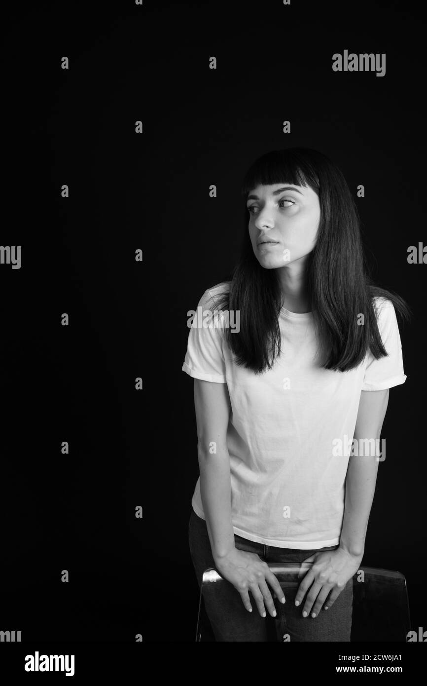 Studio Portrait Of A Pretty Brunette Woman In A White Blank T Shirt Standing Behind A Chair Against A Plain Black Background Looking To The Side Stock Photo Alamy