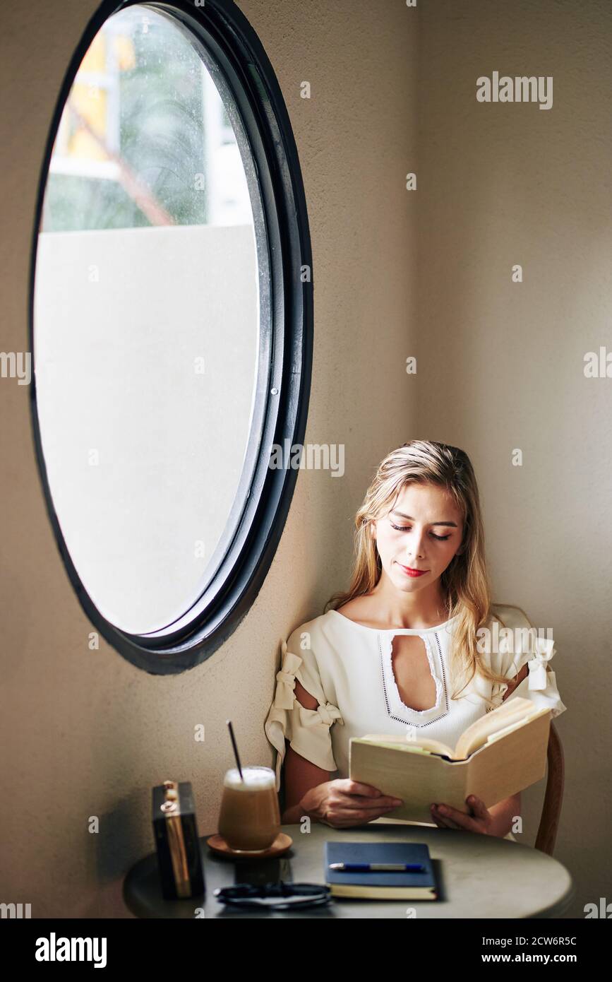 Charming smiling young woman sitting at table and reading interesting novel Stock Photo