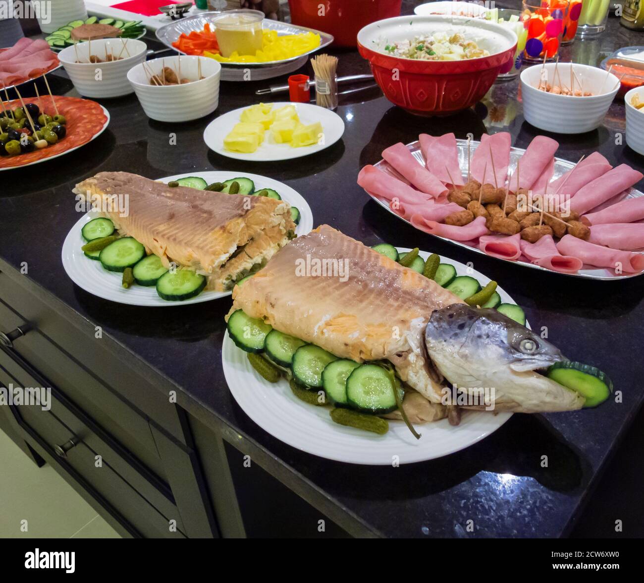 A buffet of mixed cold food laid out in a kitchen prepared for a party, including a whole salmon with cucumber slices, cold meats and sausages Stock Photo