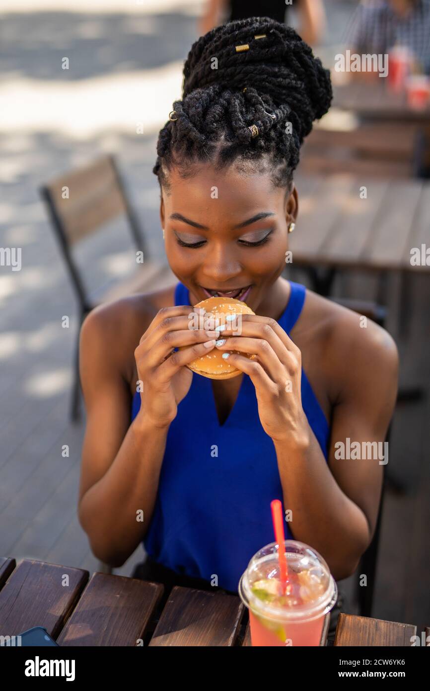 Happy African tourist having snack in burger bar, while using laptop to find out new location to reach. Stock Photo
