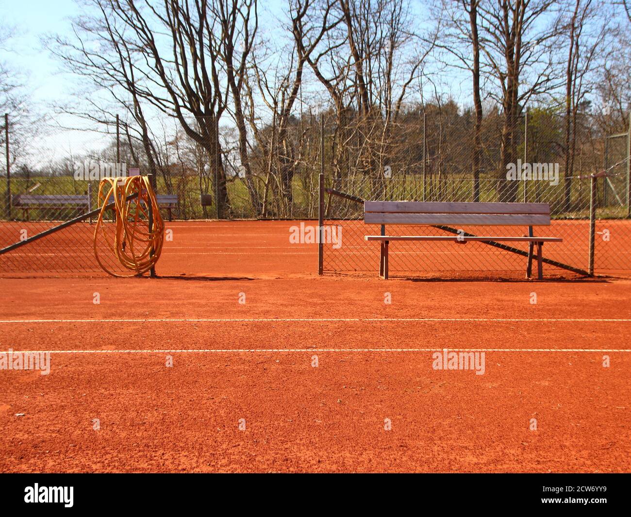 Detail of an empty red clay tennis court in the summer. There is a wooden bench and a yellow hose in the center. Stock Photo