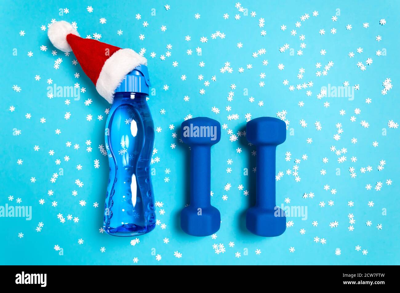 Christmas sport flat lay with dumbbells and water bottle in red Santa's hat on blue background with snowflakes. Christmas and new year holiday concept Stock Photo
