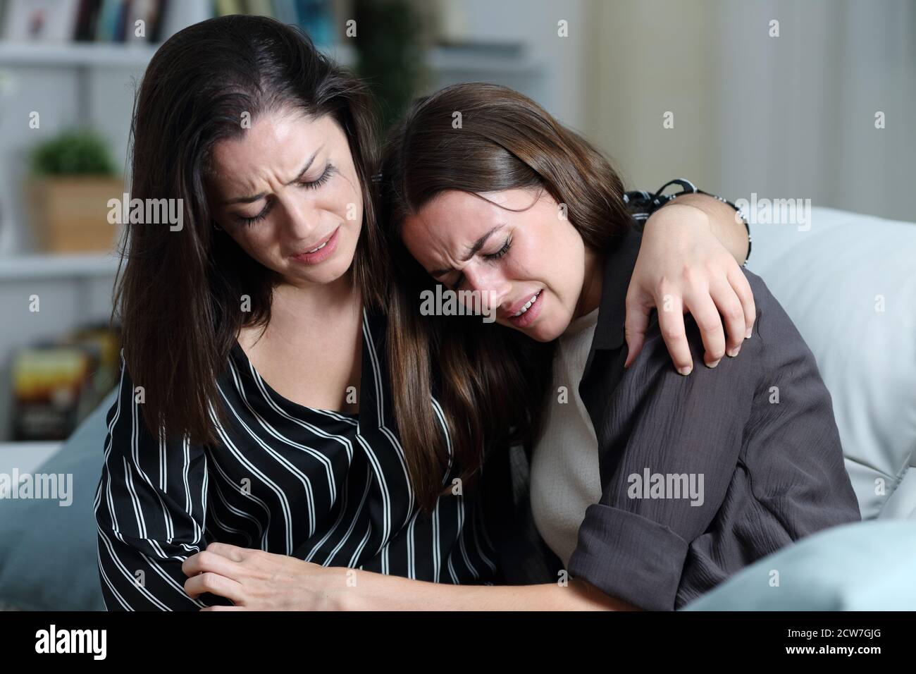 Two sad friends or sisters crying together on a couch in the living room at home Stock Photo