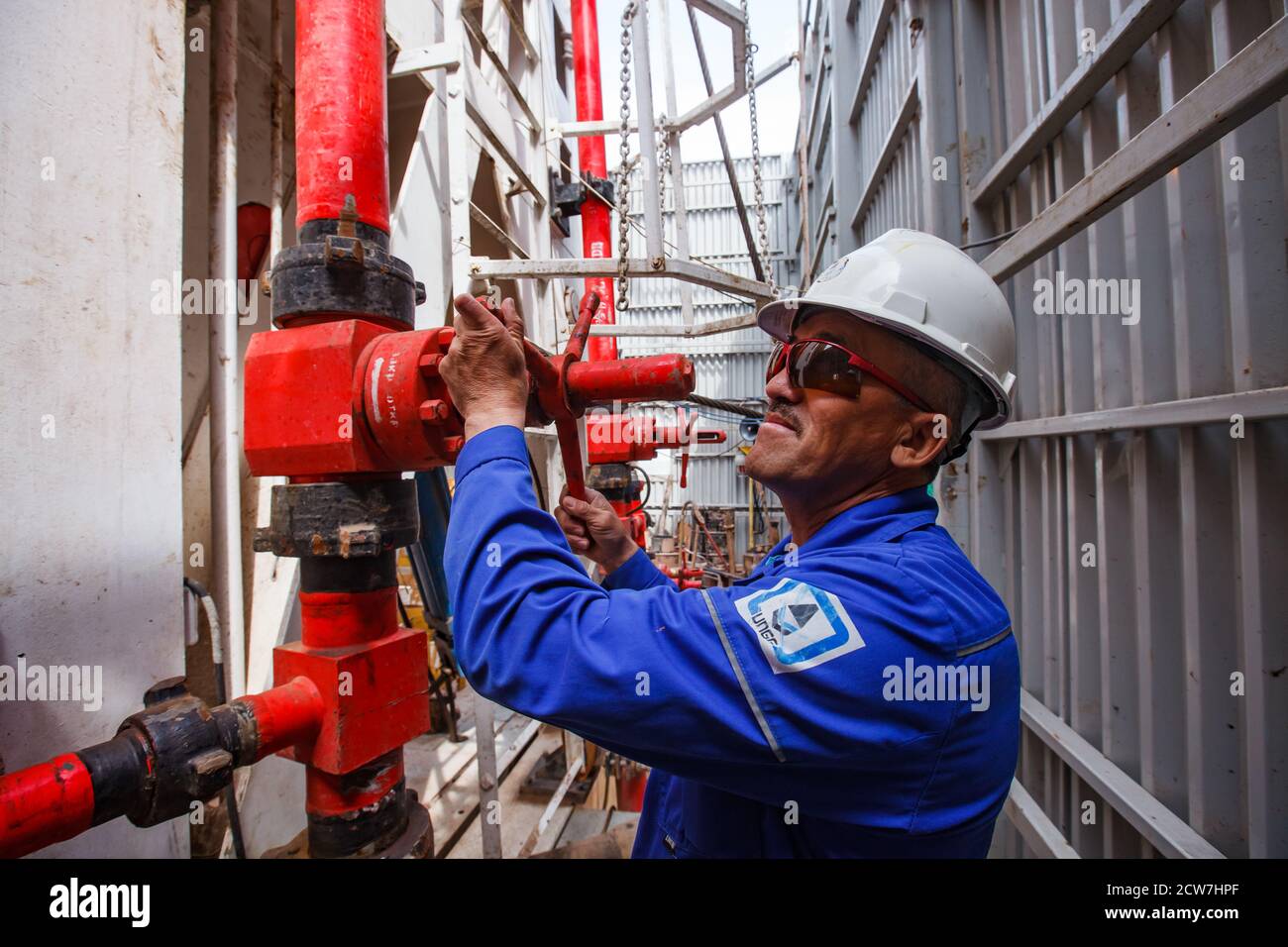 Adult asian worker in white work helmet and blue work wear adjusting valve on oil drilling rig. Zhaik-Munai oil deposit, Kazakhstan Stock Photo