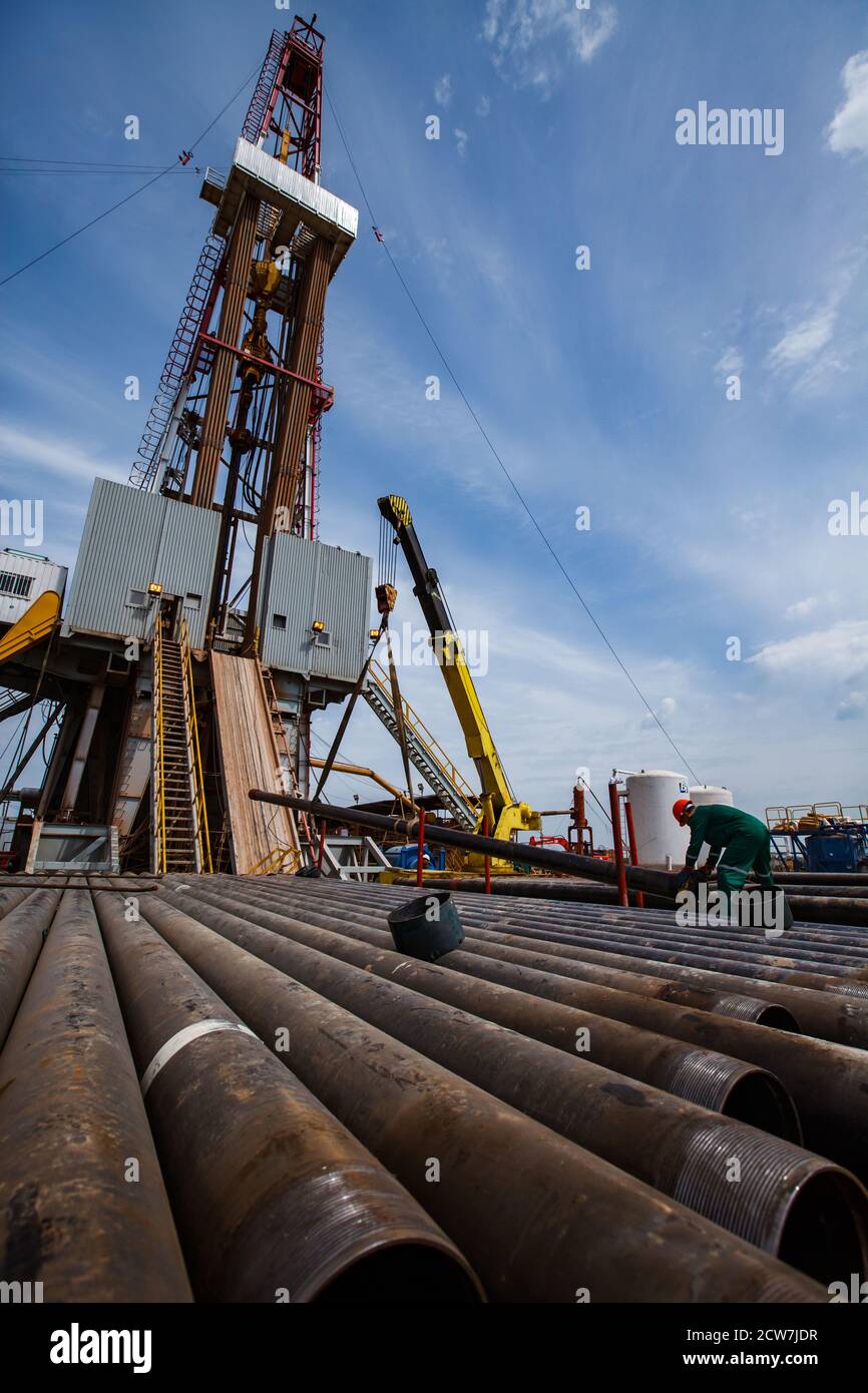 Works on drilling rig on oil deposit. Rusted drilling pipes and oil worker on foreground. Blue sky and light clouds. Wide-angle photo.. Stock Photo