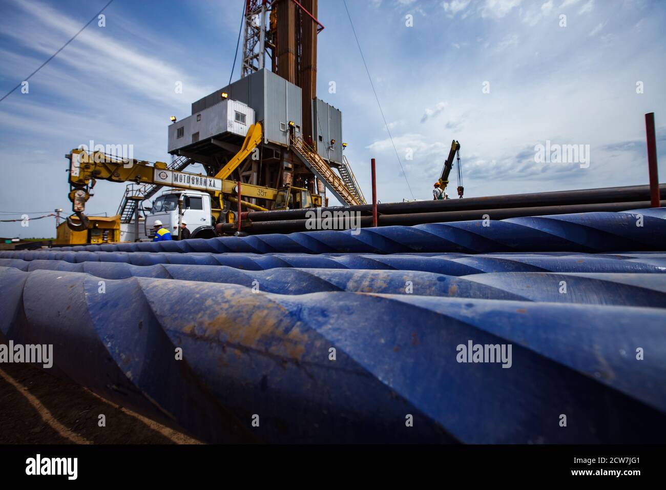 Oil deposit. Works on drilling rig. Blue rusted drilling pipes on front in focus. Oil worker, truck crane and rig on background. Blue sky and cloud. Stock Photo