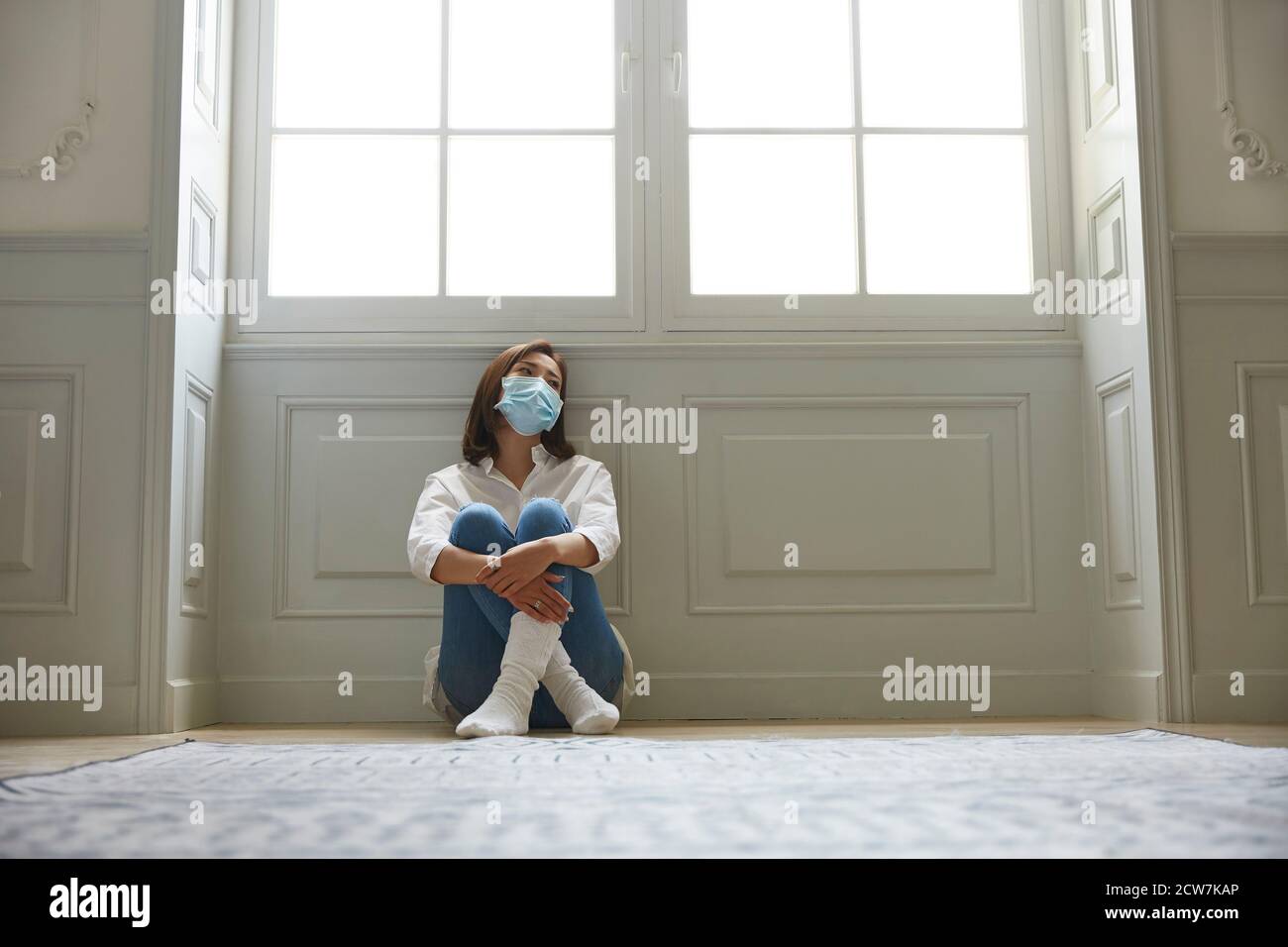 young asian woman in quarantine at home wearing facial mask sitting on floor legs crossed looking sad and depressed Stock Photo