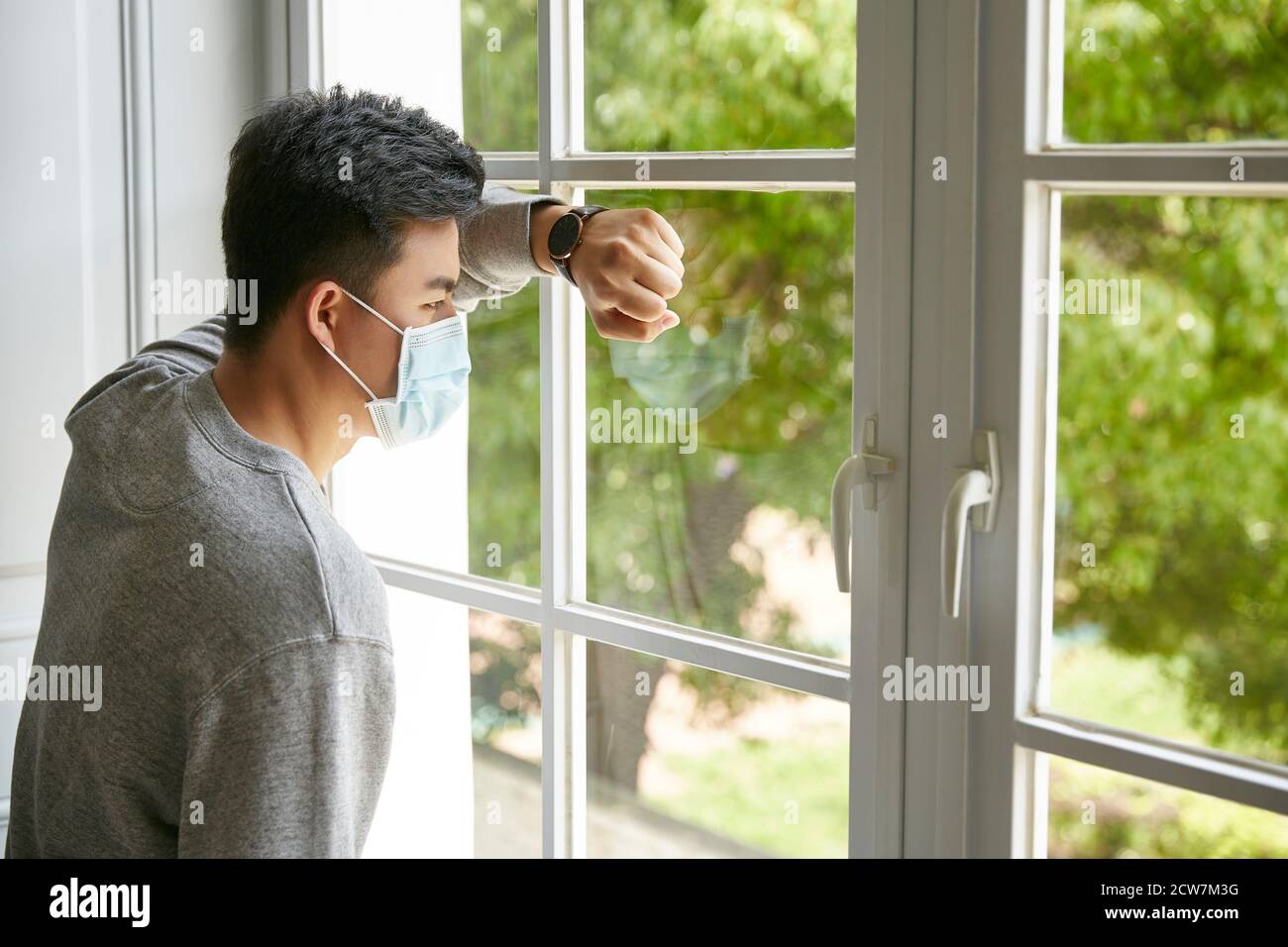 young asian man leaning against window looking at trees outside Stock Photo