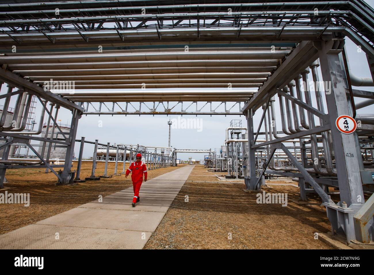 Oil worker in red work wear and red  helmet under the pipelines. Oil refinery plant in desert with yellow sand. Zhaik-Munai oil deposit, Kazakhstan. Stock Photo