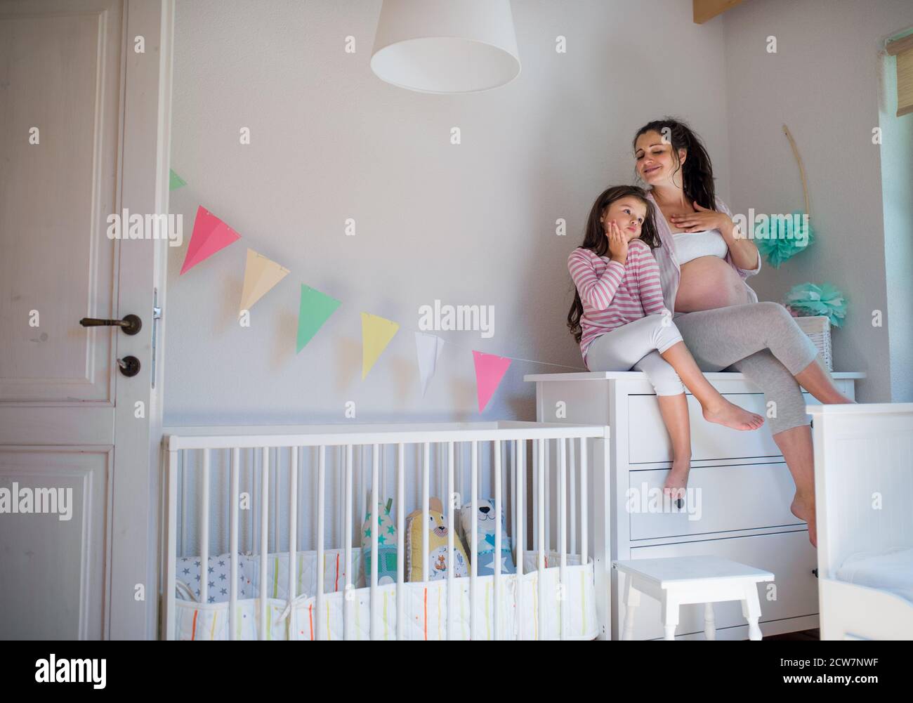 Portrait of pregnant woman with small daughter indoors at home, sitting on chest of drawers. Stock Photo