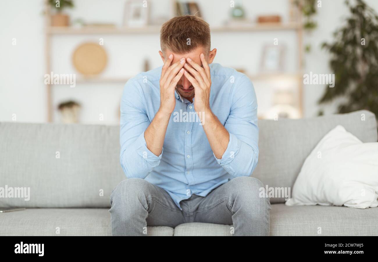 Depressed guy sitting on couch, covering face with hands Stock Photo