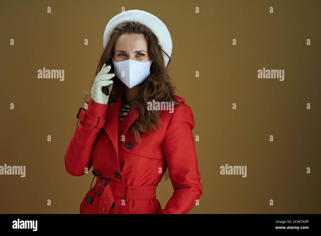 Life during covid-19 pandemic. smiling stylish woman in red coat using a smartphone against brown background. Stock Photo