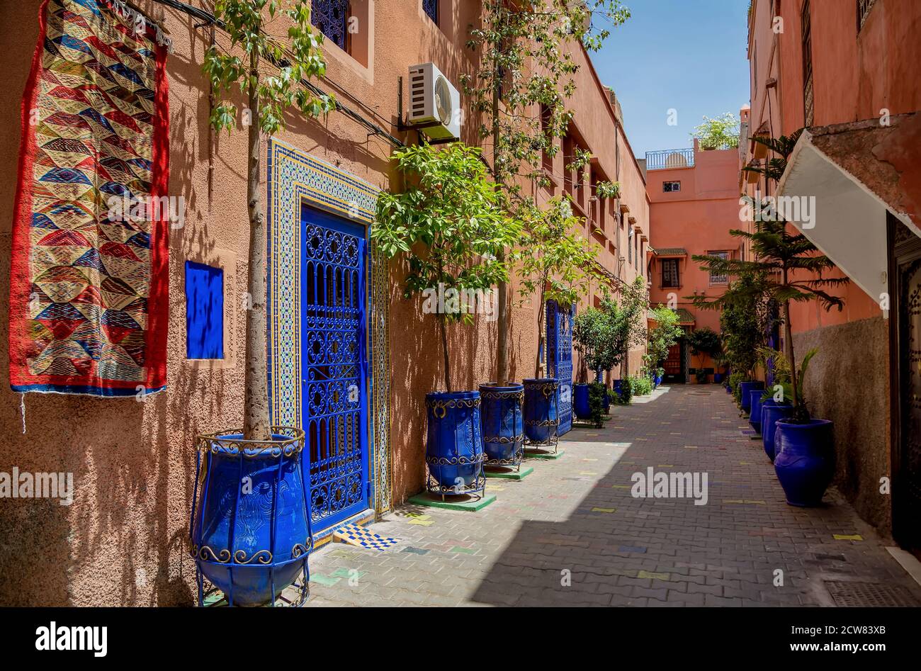 Typical narrow street in Marrakesh with carpet, bright blue ceramic flower pots, blue painted doors, red whitewashed wall. Stock Photo