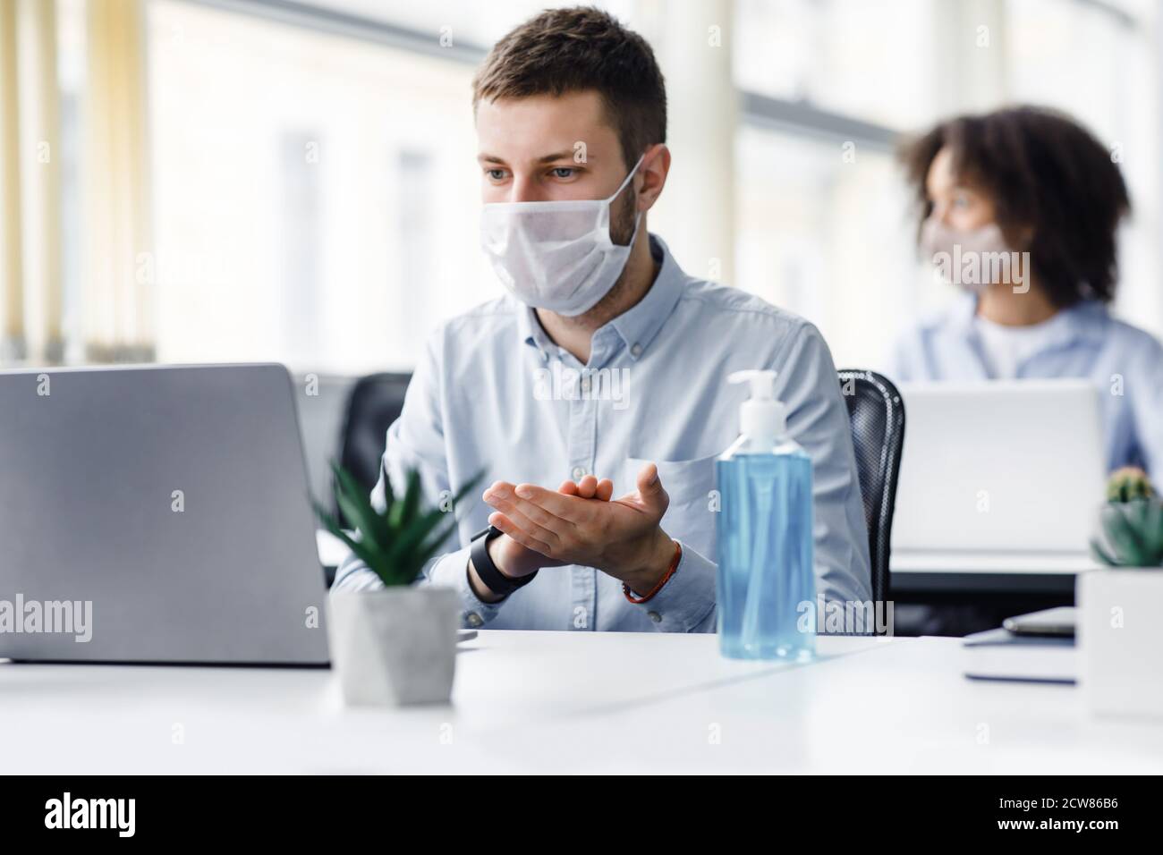 Work, new normal and health protection in workplace. Young man in protective mask disinfects hands with antiseptic and looks at laptop Stock Photo