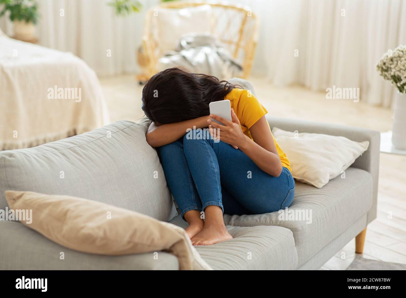 Sad young black woman with smartphone crying on sofa at home Stock Photo