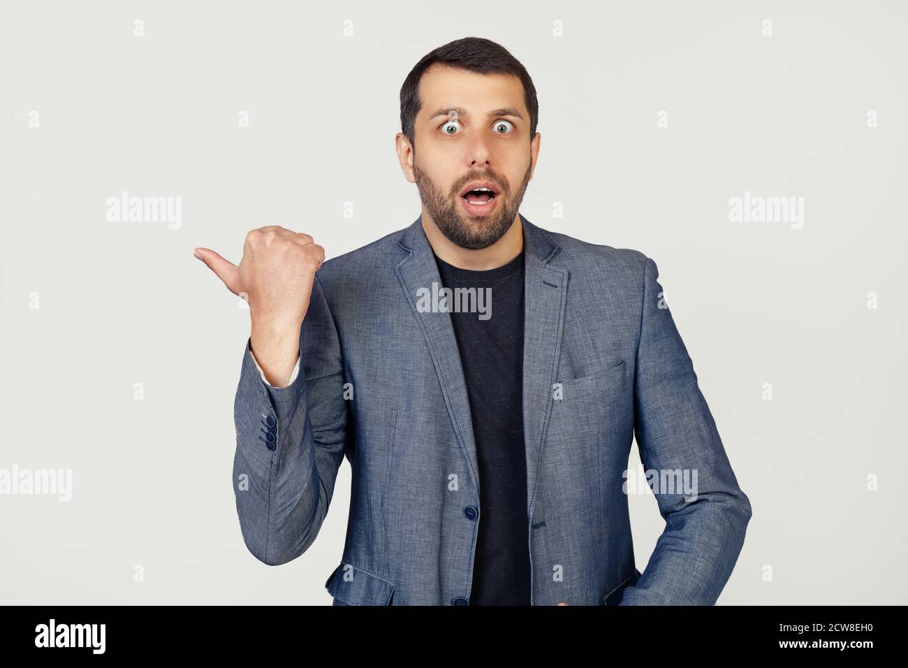Young businessman man with a beard in a jacket, surprised, pointing finger to the side, with open mouth amazed expression. Portrait of a man on a gray background. Stock Photo