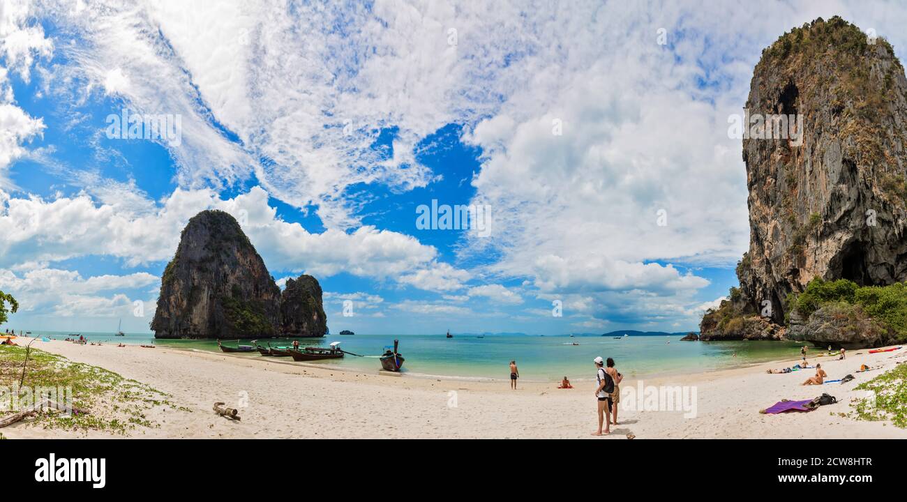 Full panorama of PhraNang Cave Beach and Thaiwand Wall in andaman sea. Sandy beach with tourists walking and swimming, long-tail boats, mountain and blue sky with clouds Stock Photo
