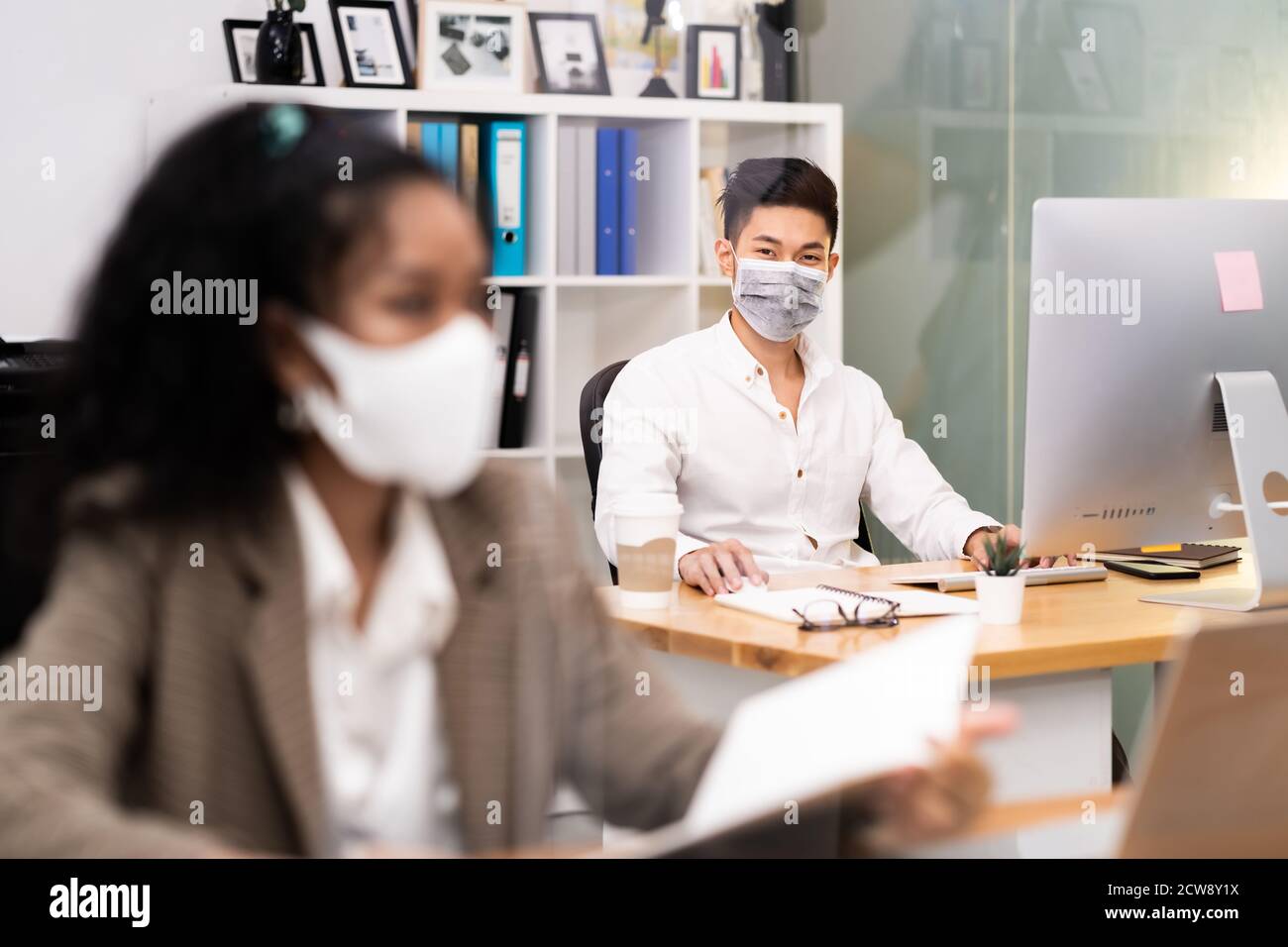 Portrait asian Office employee with protective face mask working in new normal office with social distance practice to american african colleague in f Stock Photo