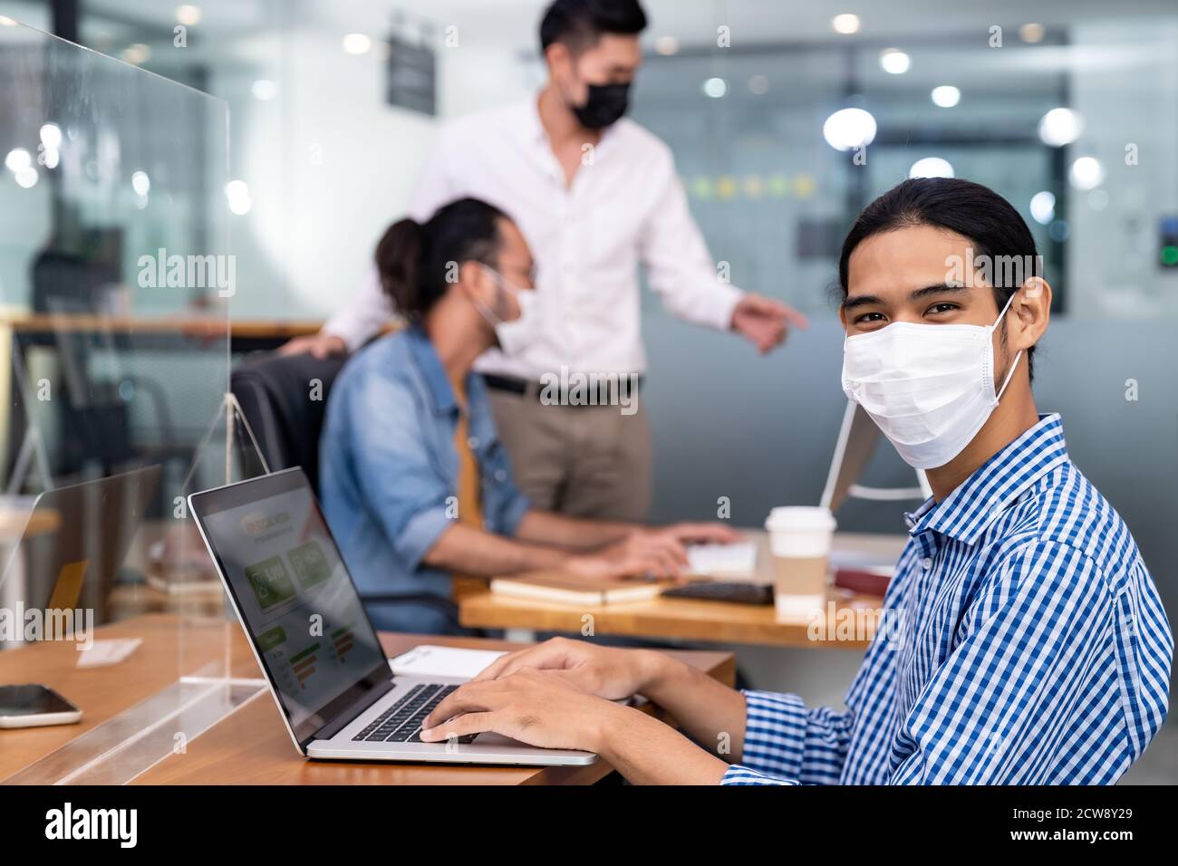 Asian Office employee with protective face mask working in new normal office with social distance practice to his colleague in background prevent coro Stock Photo