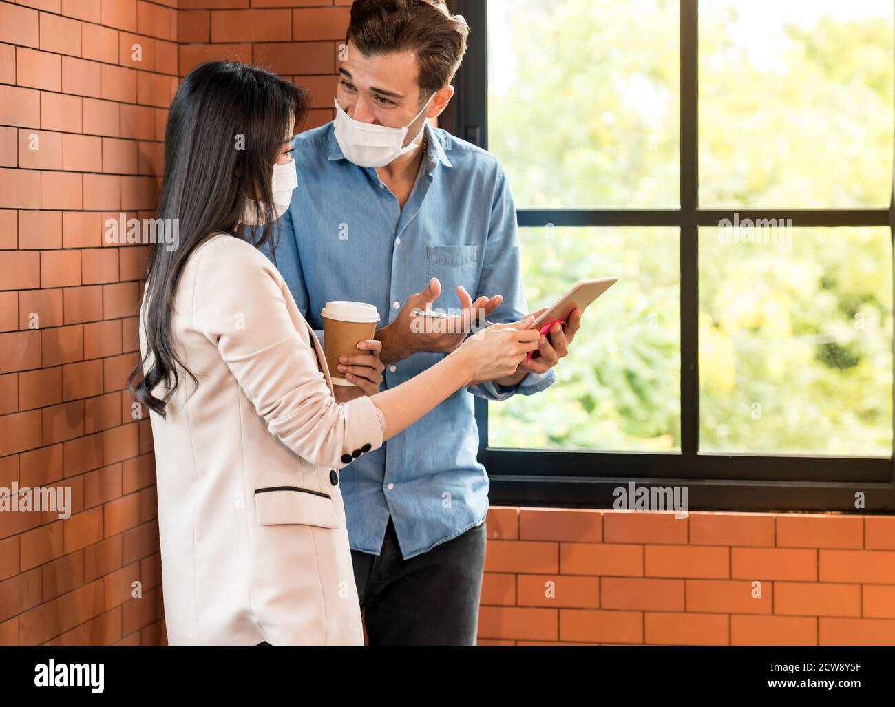 Two business person standing for brainstorm and discuss their work with digital tablet. They wear protective face mask in new normal office preventing Stock Photo
