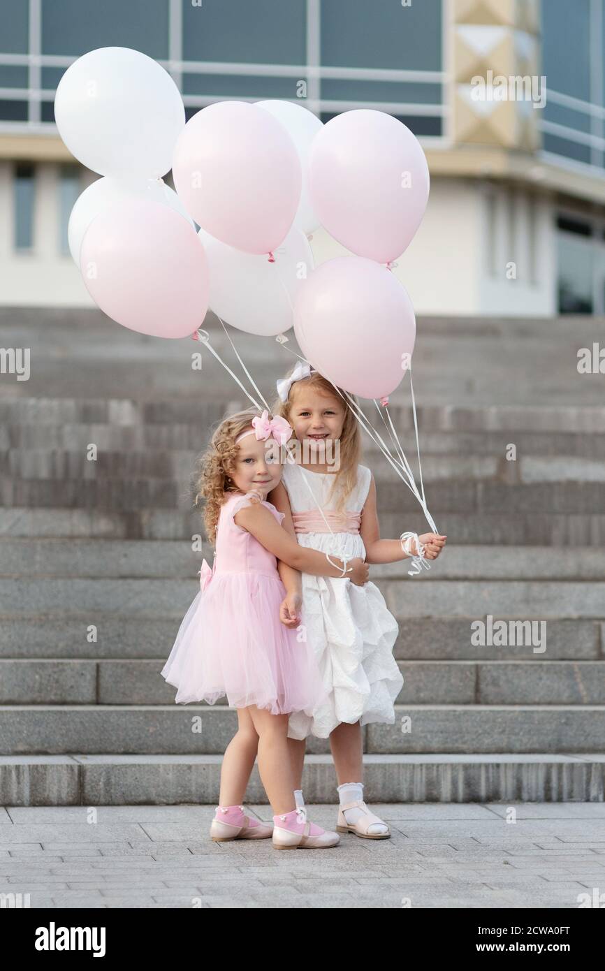 Two little girls in pink and white dressses with balloons hugging on ...