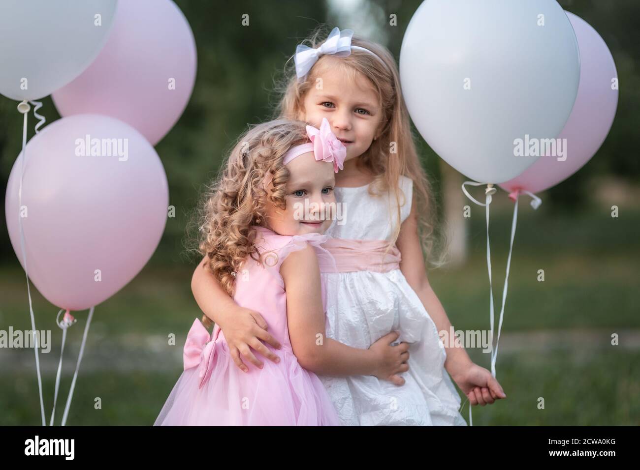 Little girls in pink and white dressses with balloons hugging in the ...